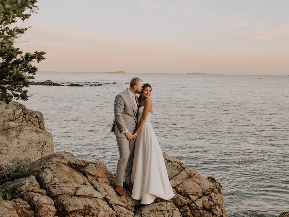 Groom in a light gray suit kissing bride's forehead on a rocky shore at ocean sunset.